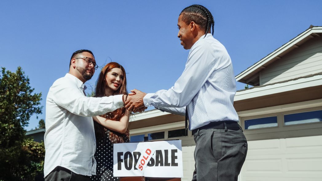 Man in white shirt wearing glasses, with woman in brown polka-dot dress and long red hair by his side, shaking hands with a man in a blue striped shirt and gray pants with dark hair, over a “Sold” sign in front of a house that was sold during the 2021 Scottsdale housing market.