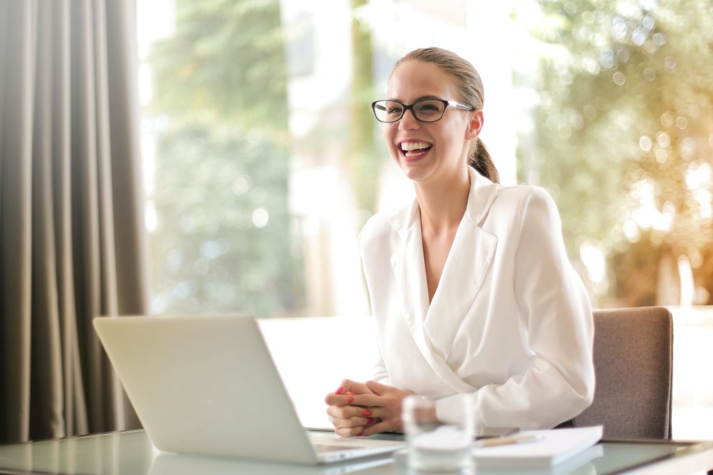 Smiling woman with brown hair pulled back into a ponytail, wearing white jacket and dark-framed glasses, looking up from a computer that is showing how KW technology ramps up her real estate business.