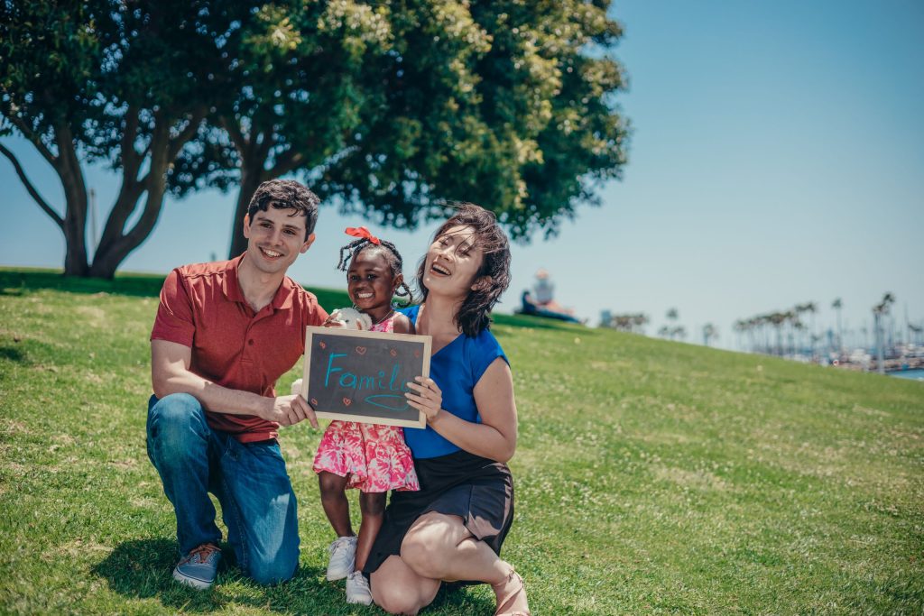 Man wearing red shirt and jean, kneeling, smiling and hold sign; woman wearing blue shirt and black skirt, kneeling, holding sign; girl wearing red dress and white sneakers, behind the sign; sign has “Family” written on it, representing Keller Williams Culture of God, Family, and then Business.