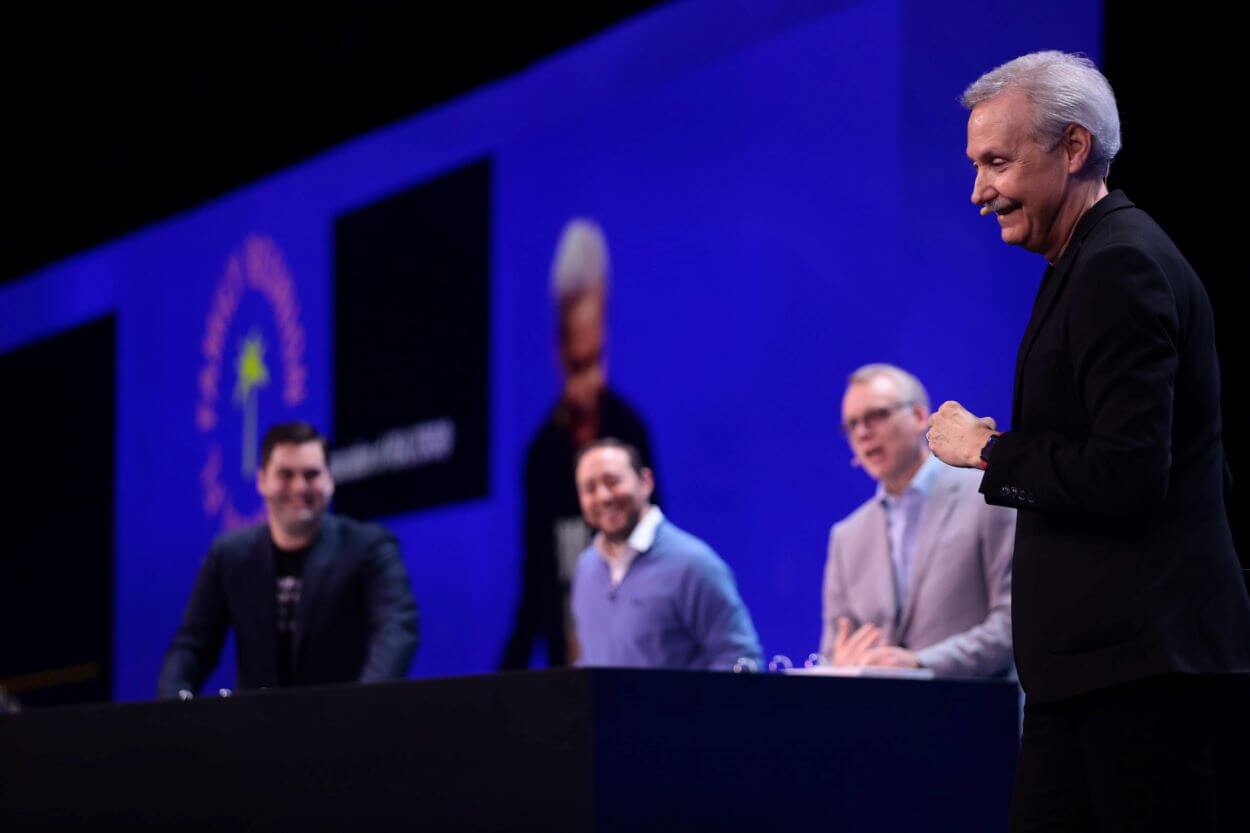 Gary Keller speaking at Family Reunion 2022 while KWAZ ranks Top 200, helping to propel the company forward, three other company leaders are seated at a table behind Gary who is wearing a dark suit, facing left, and smiling