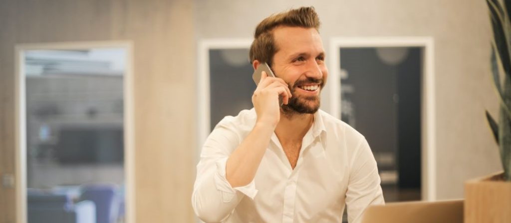 Handsome man with brown hair, mustache, and beard, with tattoos on his left hand and wrist, wearing a white shirt, talking on a cell phone and practicing the Top 5 Cold Calling Tips for real estate success.