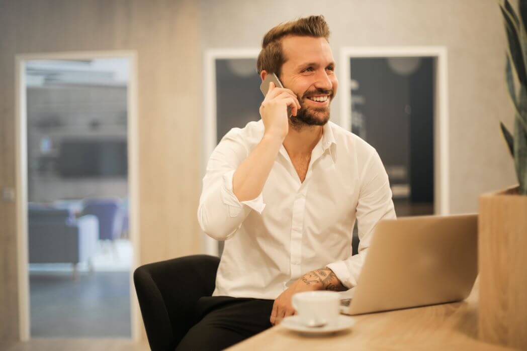 Handsome man with brown hair, mustache, and beard, with tattoos on his left hand and wrist, wearing a white shirt, talking on a cell phone and practicing the Top 5 Cold Calling Tips for real estate success.