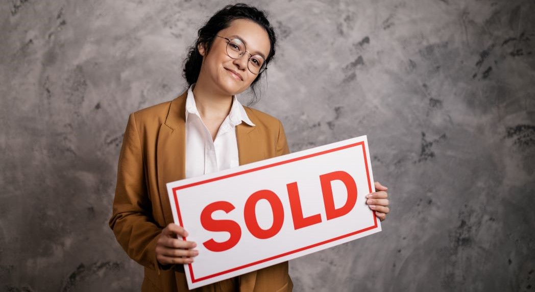 Woman real estate agent wearing brown blazer and white shirt holding sign with SOLD in red letters, and she is ready to cap. She is wearing round wire-frame glasses and has black shoulder length hair, facing forward and smiling.
