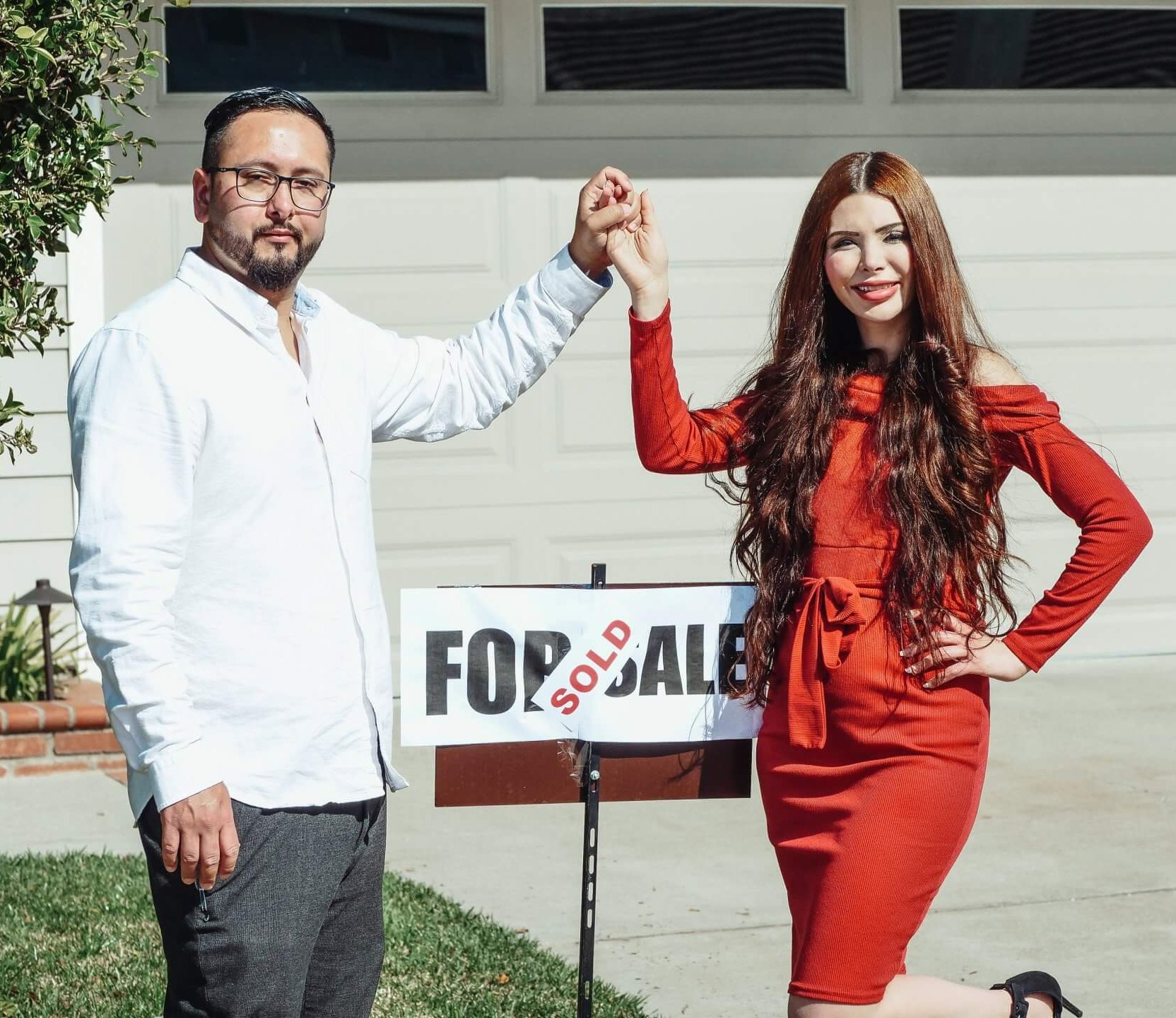 Man with glasses and white shirt holding hands with woman who is wearing a red dress over a sold sign in front of a house they purchased knowing the top 5 reasons to buy a home worked best for their buying situation.